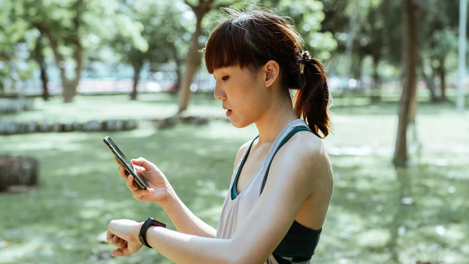 A runner in a park checking her phone in one hand and her watch on the other wrist.