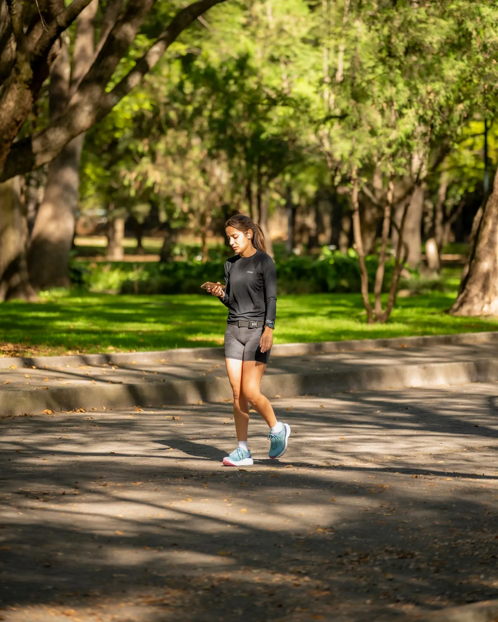 A runner walking a tree-lined park path, looking at her phone.