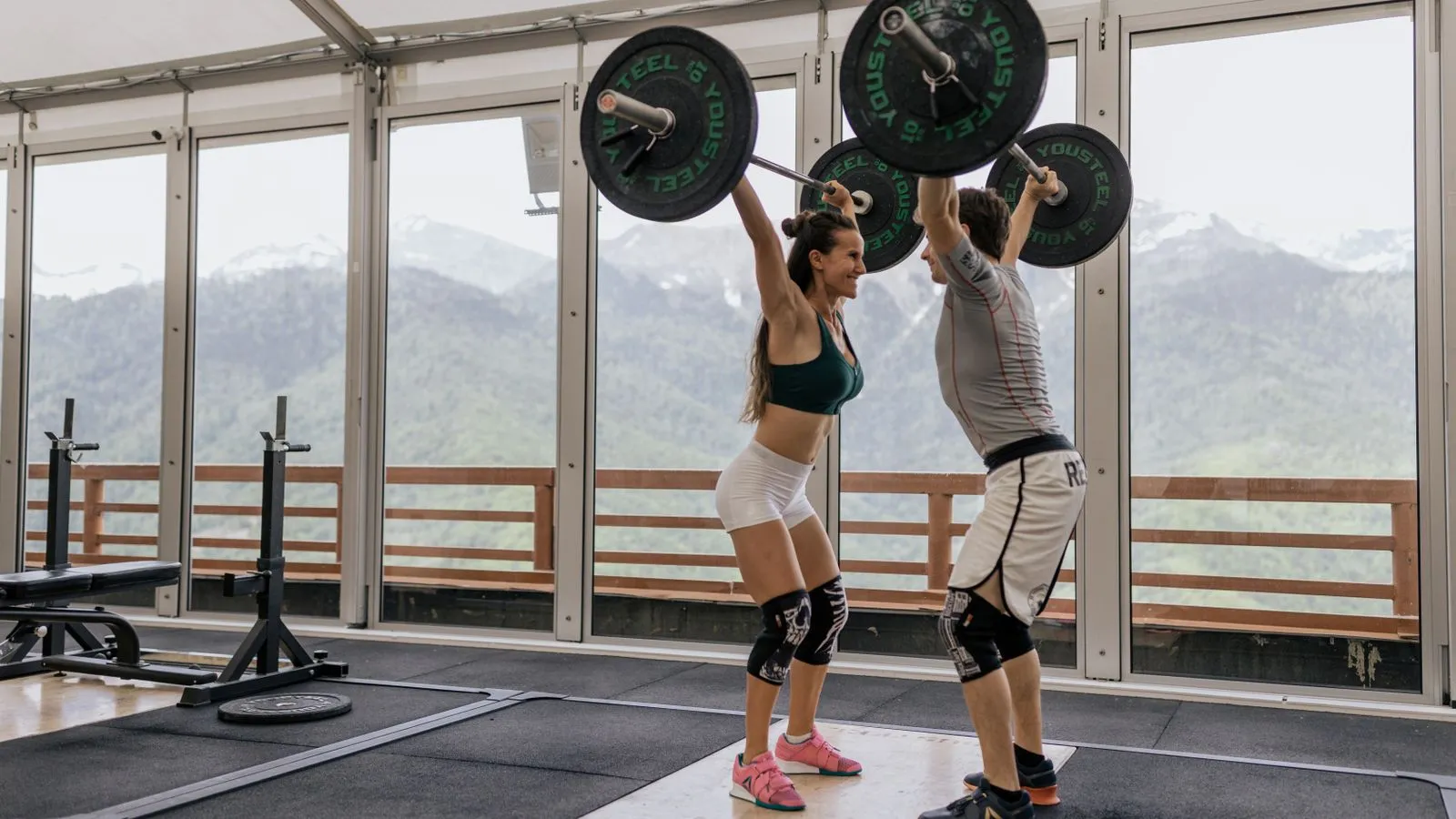 Two lifters pressing barbells overhead in a gym with a wall of windows looking out at snow-capped mountains.