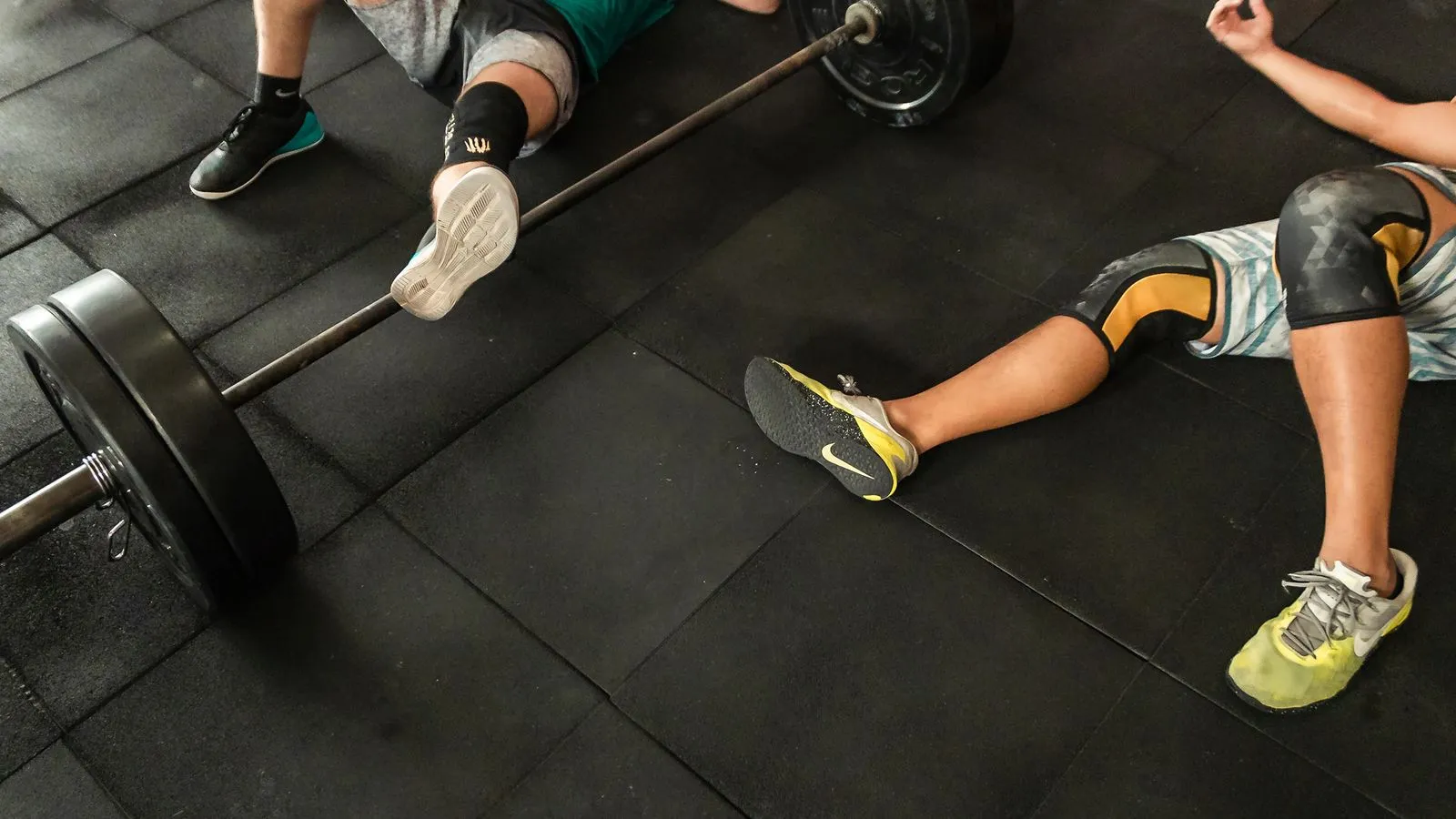 Two lifters sprawled on a gym floor next to a loaded barbell, resting between sets.
