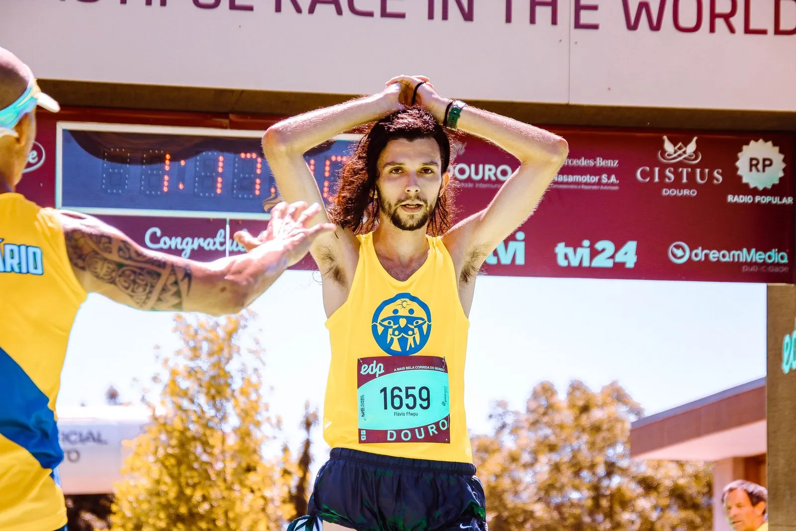 A runner crossing a race finish line with his hands on his head, the clock still running behind him.
