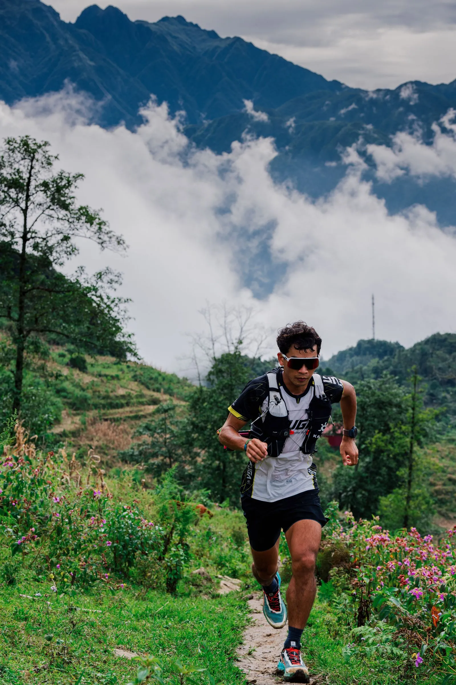 A trail runner in a hydration vest climbing a narrow path, with cloud sitting below the mountains behind him.