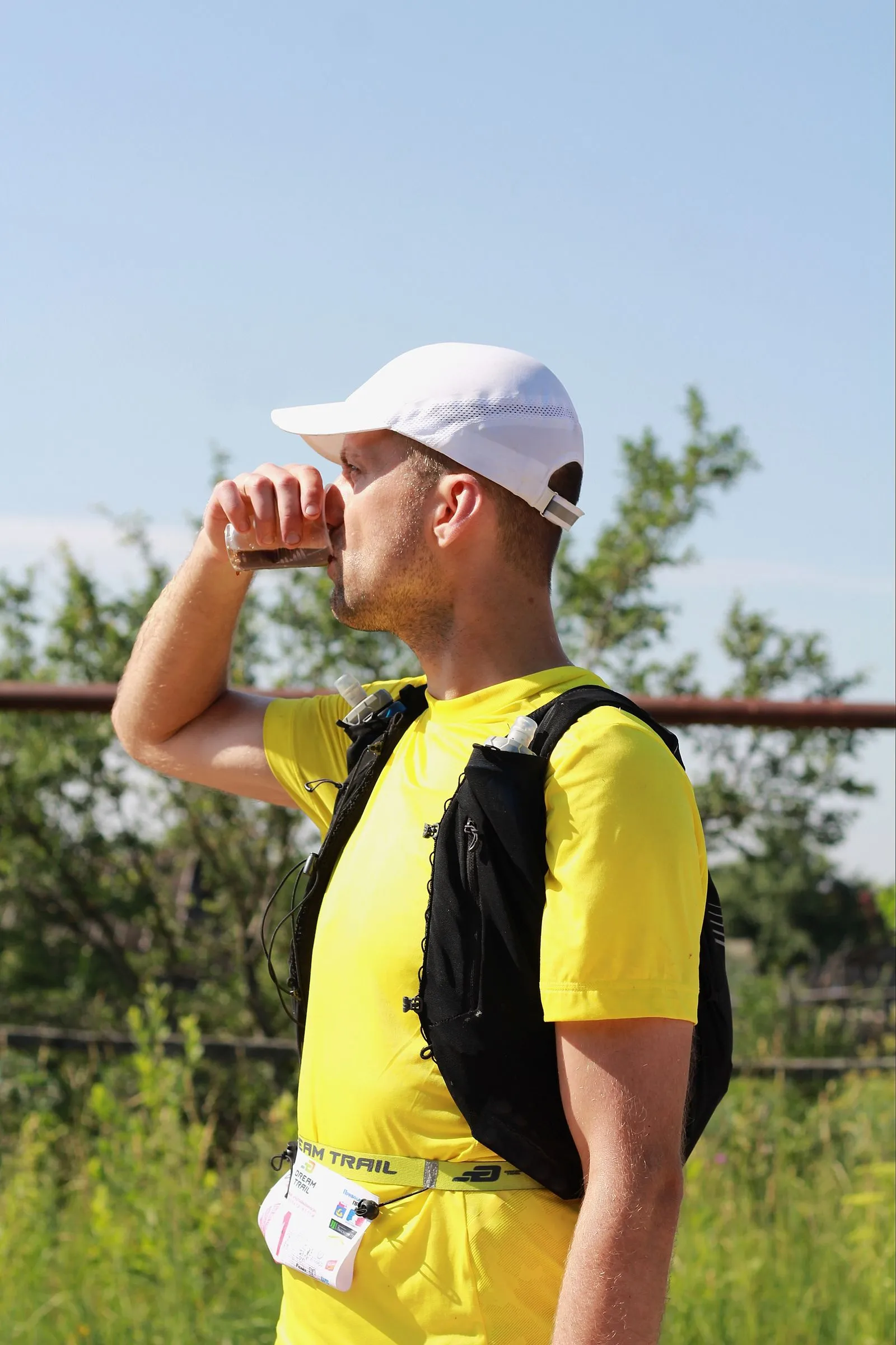 A trail runner in a hydration vest drinking from a cup at an aid station.