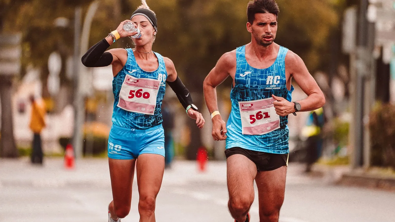 Two race runners side by side on a road, one drinking from a water bottle mid-stride.